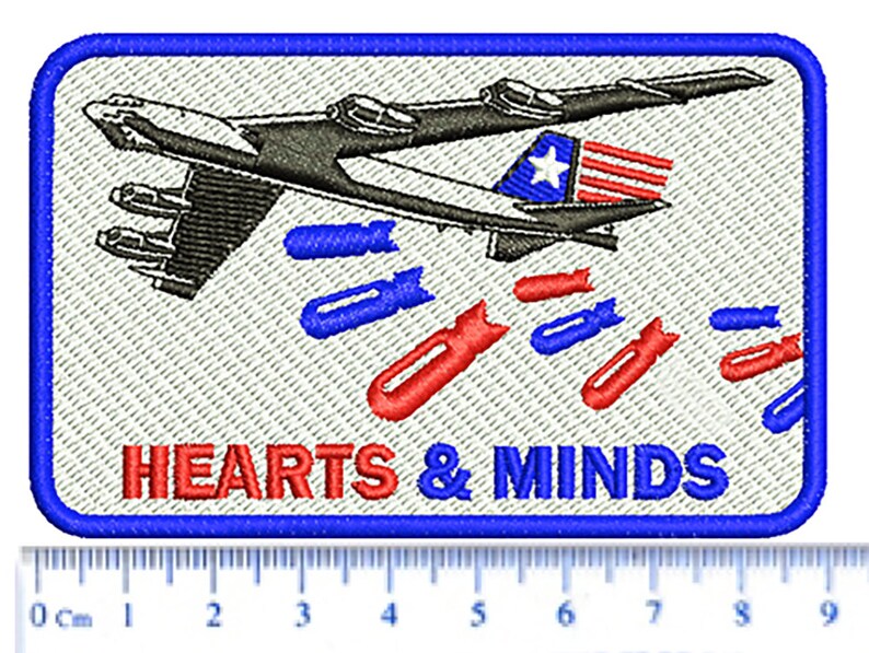 以下が含まれることがあります： 青い縁取りのある白い刺繍パッチ。パッチには、爆撃機が爆弾を投下している白黒のイラストが描かれています。爆弾は赤、白、青です。飛行機の下には「HEARTS & MINDS」という文字が赤と青の文字で書かれています。