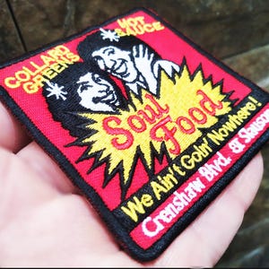 Puede incluir: Un parche cuadrado bordado con fondo rojo y borde negro. El parche presenta las palabras "Soul Food" en rojo y amarillo, con ilustraciones de caras sonrientes. El texto incluye "Collard Greens" y "We Ain't Goin' Nowhere!"