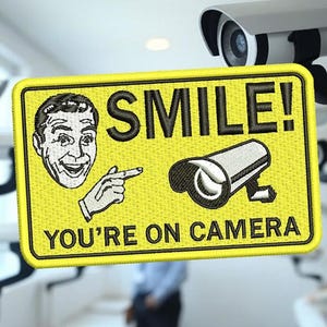May include: Yellow patch with a black border, displaying the text "SMILE! YOU'RE ON CAMERA". The patch features a smiling face, a pointing hand, and a security camera illustration. Security cameras are visible in the background.