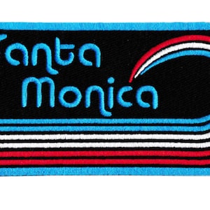 Puede incluir: Parche bordado con el texto "Santa Monica" en azul. El parche presenta un fondo negro con rayas azules, blancas y rojas y un borde azul. Un dise&ntilde;o de estilo retro.