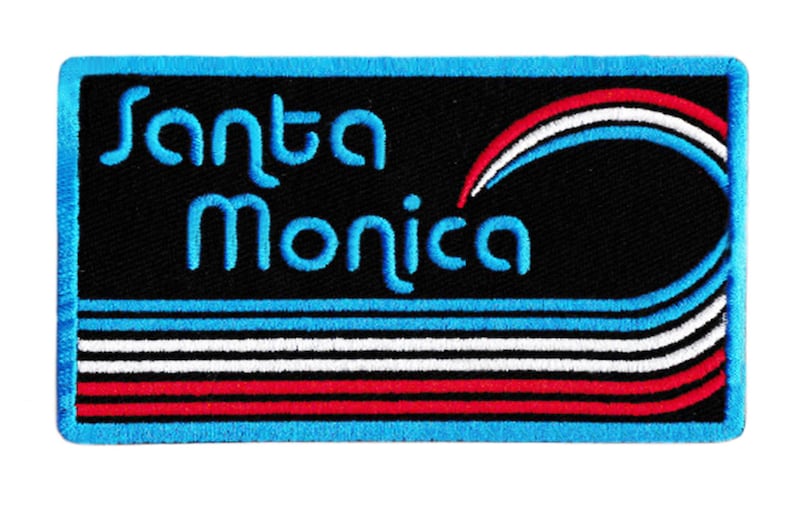 Puede incluir: Parche bordado con el texto "Santa Monica" en azul. El parche presenta un fondo negro con rayas azules, blancas y rojas y un borde azul. Un dise&ntilde;o de estilo retro.