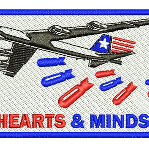 以下が含まれることがあります： 青い縁取りのある白いパッチには、赤と青の爆弾を投下する爆撃機の白黒イラストが描かれています。飛行機の下には「HEARTS & MINDS」という文字が赤と青の文字で印刷されています。