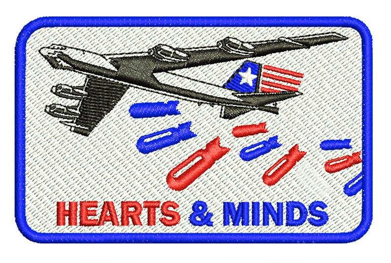 以下が含まれることがあります： 青い縁取りのある白いパッチには、赤と青の爆弾を投下する爆撃機の白黒イラストが描かれています。飛行機の下には「HEARTS & MINDS」という文字が赤と青の文字で印刷されています。