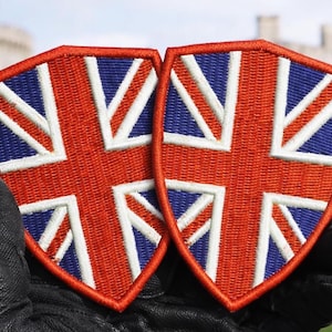 May include: Two shield-shaped patches featuring the Union Jack flag design. The patches have a red border and background, with white and blue accents. The patches are held by a person wearing a black glove.