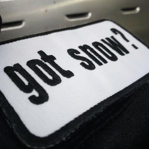 May include: A white rectangular patch with black embroidered text that reads "got snow?". The patch has a black border and is set against a dark background. The patch is likely for a jacket or bag.