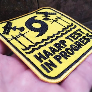 May include: Yellow patch with black text and a black image of a hurricane. The text reads "HAARP TEST IN PROGRESS".