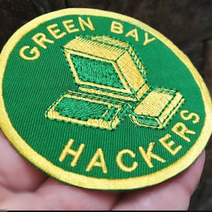 Może przedstawiać: Okrągła, haftowana naszywka z zielonym tłem i żółtą obwódką. Naszywka przedstawia grafikę komputerową i słowa "GREEN BAY HACKERS" w żółtych literach. Naszywka jest okrągła i wykonana z tkaniny.