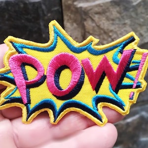 Może przedstawiać: Naszywka haftowana na żółto, niebiesko i różowo z napisem "POW!" w stylu komiksowym.
