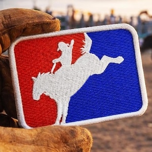 May include: A rectangular patch with a white embroidered image of a rodeo rider on a bucking bronco. The patch is divided into red and blue sections, with a white border. The patch is held by a brown leather glove.
