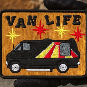 May include: Embroidered patch with the words "VAN LIFE" in black letters above a black van with a red, yellow, and white design. The patch has a gold border and is held by a gloved hand.
