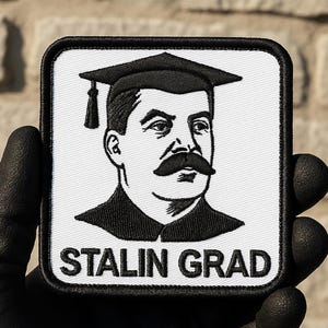 Puede incluir: Parche cuadrado con borde negro y fondo blanco. El parche presenta una ilustración en blanco y negro de un hombre con birrete y un gran bigote. El texto "STALIN GRAD" está impreso en negro en la parte inferior del parche.