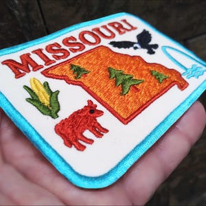 以下が含まれることがあります： 赤い「MISSOURI」の文字、オレンジ色の州の輪郭、緑の木、赤い牛、トウモロコシの穂、黒い鳥、滝が刺繍されたパッチ。 パッチは白い背景と水色の縁取りです。