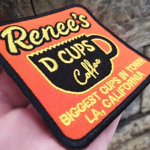 Può includere: Toppa ricamata con la scritta "Renee's D Cups Coffee" in giallo su sfondo arancione. Al centro è presente una grafica di tazza di caffè nera. È incluso anche il testo "Biggest Cups in Town LA, California".