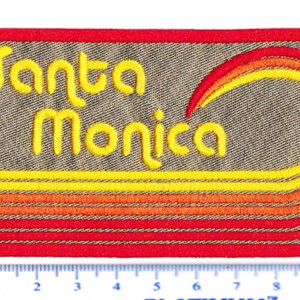 Puede incluir: Parche bordado con las palabras "Santa Monica" en escritura amarilla. El parche presenta un dise&ntilde;o de l&iacute;neas horizontales amarillas, naranjas y rojas y un arco&iacute;ris curvo. El parche es rectangular con un borde rojo.