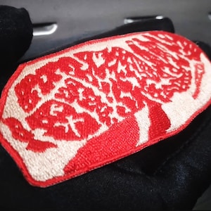 May include: A red and white embroidered patch with a detailed design. The patch is held in a black gloved hand. The design features a red border and a white pattern with Japanese characters.