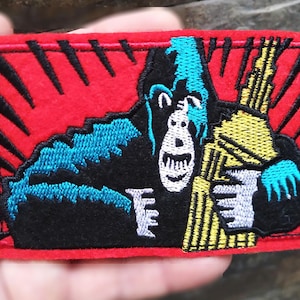 Peut inclure: Un écusson en feutre rouge avec un contour noir représente un gorille bleu et noir tenant un objet rayé jaune et blanc. Le gorille a un visage blanc avec une bouche et des yeux noirs. L'arrière-plan est rouge avec des rayons noirs qui rayonnent du centre.