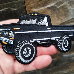 Puede incluir: Parche bordado negro de una camioneta vintage con contorno blanco. La camioneta tiene neumáticos grandes y una caja negra.