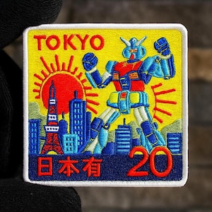Puede incluir: Parche bordado con un robot frente al horizonte de Tokio con un sol rojo. El parche tiene la palabra "TOKYO" en rojo, caracteres japoneses y el número "20". El fondo es amarillo.