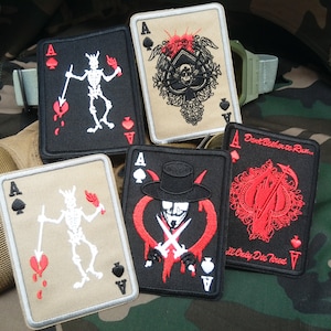May include: Four embroidered patches featuring the Ace of Spades with different designs. The patches are black and beige with red accents. The designs include a skeleton holding a flaming torch, a skull with crossed swords, a masked figure with crossed swords, and a scythe with a red heart.