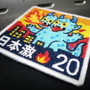 Könnte beinhalten: Gestickter Aufnäher mit zwei blauen Cartoon-Monstern mit Hörnern und roten Zungen, vor einem feurigen Hintergrund. Der Aufnäher enthält japanischen Text und die Zahl 20, mit einem weißen Rand.