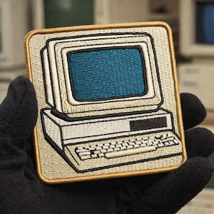 Pode incluir: Patch bordado com um design de computador retro. O patch tem uma borda bege e mostra um computador vintage com tela azul, caixa bege e teclado. O design é delineado com fio preto.