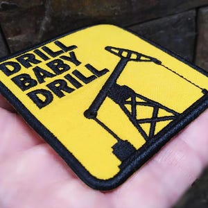 Puede incluir: Parche bordado amarillo con costuras negras. El parche presenta una torre de perforación de petróleo y el texto "DRILL BABY DRILL".
