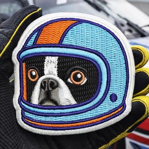 Op de afbeelding: Geborduurde patch met een Boston Terrier-hond met een racehelm. De helm is lichtblauw met oranje en donkerblauwe accenten. Het gezicht van de hond is zwart en wit met bruine ogen. De patch wordt vastgehouden door een gehandschoende hand.
