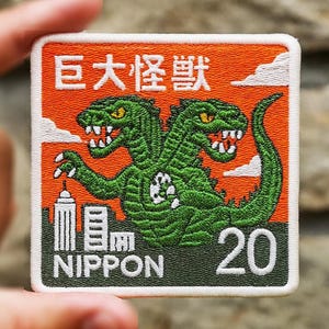 May include: Embroidered patch featuring a two-headed green monster with yellow eyes, set against an orange background. The patch includes Japanese text, the word "NIPPON", and the number "20". The design is framed by a white border.