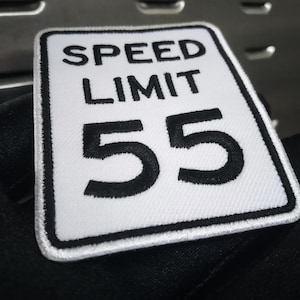 May include: White rectangular patch with black embroidered text that reads "SPEED LIMIT 55". The patch has a black border and is held against a dark background. The patch is likely an accessory.