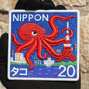 May include: Embroidered patch with a blue background featuring a red octopus, Tokyo Tower, and the word "NIPPON". The patch also includes Japanese text and the number 20. The patch is square with a white border.