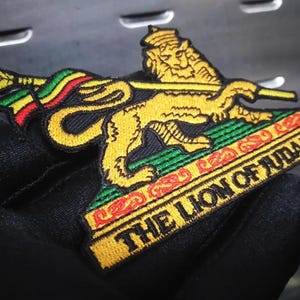 May include: Embroidered patch featuring a yellow lion holding a staff with red, yellow, and green stripes. The patch has the text "THE LION OF JUDAH" in yellow on a black background, with a green and red border.