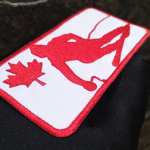 May include: Embroidered patch with a red climber silhouette and maple leaf on a white background, framed by a red border. The patch has a rectangular shape.