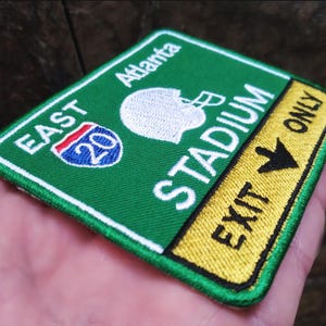 May include: Embroidered patch with a green background, white text, and a football helmet graphic. The patch reads "East Stadium" and "Exit Only" with an Interstate 20 sign. The patch is rectangular with a white border.