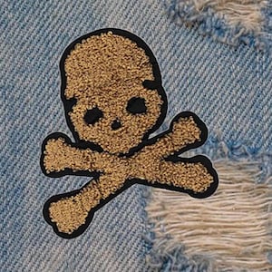 Könnte beinhalten: Ein gestickter Totenkopf- und Knochen-Patch in Braun und Schwarz. Der Totenkopf ist flauschig und die Knochen sind mit einem schwarzen Umriss gestickt.
