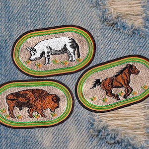 May include: Three embroidered patches on a blue denim background. The patches are oval shaped and feature a white pig, a brown horse, and a brown bison. Each patch has a green and orange border.