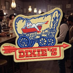 May include: A vintage-style embroidered patch featuring a red, white, and blue wagon with a woman riding inside. The wagon has blue wheels and is pulled by a red arrow. The text "DIXIE'S" is embroidered in red on a beige background.