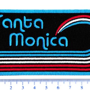 Puede incluir: Parche bordado con la frase "Santa Monica" en azul claro. El parche tiene un fondo negro con l&iacute;neas curvas azules, blancas y rojas. El borde es azul claro.