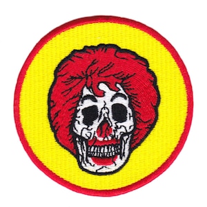 Könnte beinhalten: Ein runder, gestickter Aufnäher in Gelb und Rot mit einem Schädel, der eine rote Perücke trägt. Der Schädel hat ein weißes Gesicht mit schwarzen Augen und einem roten Mund.