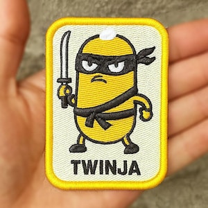 May include: Embroidered patch showing a yellow cartoon ninja character with a sword. The character wears a black eye mask and belt. The rectangular patch has a yellow border and the word "TWINJA" at the bottom, all on a cream background.