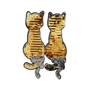 Puede incluir: Dos parches de gato de lentejuelas doradas, uno frente al otro, con contornos negros. Los gatos están sentados con las colas enroscadas detrás de ellos.