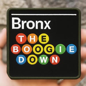 May include: A square patch with the words "Bronx THE BOOGIE DOWN" in white and colorful lettering. The patch has a black background and a green border. The patch is held in a hand.