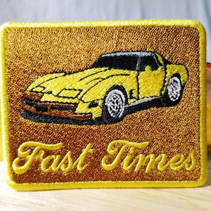Puede incluir: Parche bordado amarillo con un coche amarillo y el texto "Fast Times" en una fuente de letra dorada.