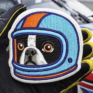 Op de afbeelding: Geborduurde patch met een Boston Terrier hond met een racehelm. De helm is lichtblauw met oranje en donkerblauwe accenten. Het gezicht van de hond is zwart en wit met grote ogen. De patch wordt vastgehouden door een gehandschoende hand.