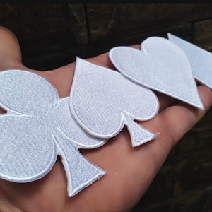 May include: White embroidered patches in the shapes of playing card suits: clubs, spades, hearts, and diamonds. The patches have a textured appearance and are held in a hand.