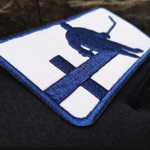 May include: Embroidered patch with a white background and a blue border. The patch features a blue silhouette of a bird perched on a cross. The patch is rectangular and appears to be sewn onto a black fabric.