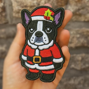 Op de afbeelding: Geborduurde patch met een Franse Bulldog verkleed als kerstman. De hond heeft een zwart-wit gezicht, draagt een rode kerstmuts met hulst. Het kerstmanpak is rood met witte randen en een gouden gesp.