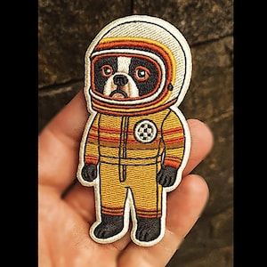 Op de afbeelding: Geborduurde patch met een Boston Terrier-hond in een astronautenpak. De patch heeft een wit, geel, oranje en zwart kleurenschema. De hond draagt een helm en een gele overall met zwarte handschoenen en laarzen.