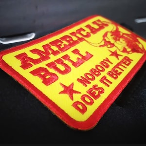Pode incluir: Patch bordado com as palavras "AMERICAN BULL" em vermelho, uma ilustração de touro e o texto "NOBODY DOES IT BETTER" sobre um fundo amarelo. O patch tem uma borda vermelha.