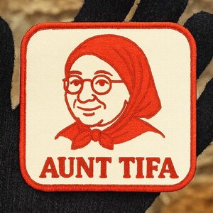 May include: Square patch with a red and white design. The patch features a cartoon illustration of a woman wearing glasses and a headscarf. The text "AUNT TIFA" is printed in red letters at the bottom of the patch.