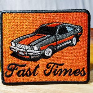 Puede incluir: Un parche bordado naranja con un coche negro y el texto "Fast Times" en letra cursiva negra.